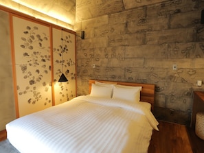 A bedroom with a bed, wooden headboard, and a stone wall with a leaf pattern. - Kawasemi no su (Nago)