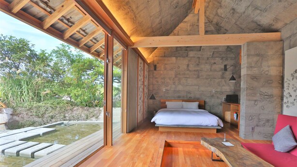 Un dormitorio moderno con suelo de madera, una cama con ropa blanca, un banco y vistas a zonas verdes.