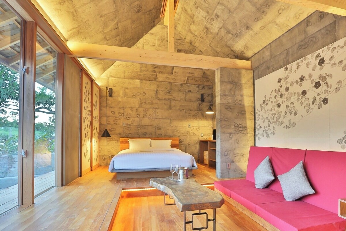 A modern bedroom with a large bed, a wooden bench, a red sofa, and a large window.