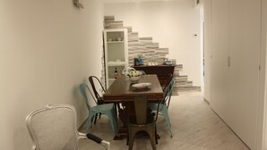 Un coin repas avec une table en bois, des chaises de couleur turquoise et une chaise blanche. Un escalier, dont la paroi est ornée d’un motif, mène à une autre pièce.