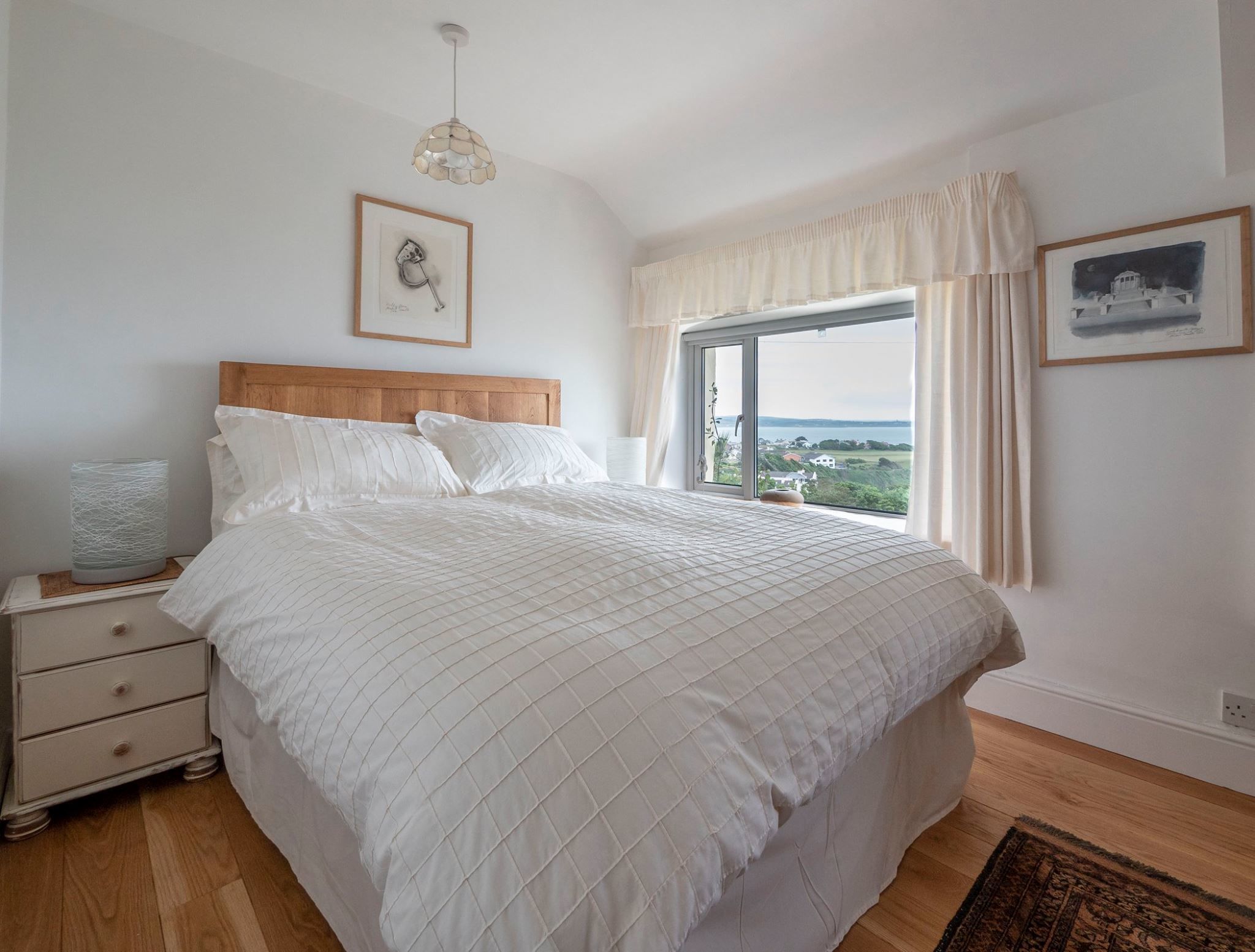 A bedroom with a large bed, a nightstand, a bedside lamp, a window with curtains, and a view of the outdoors.