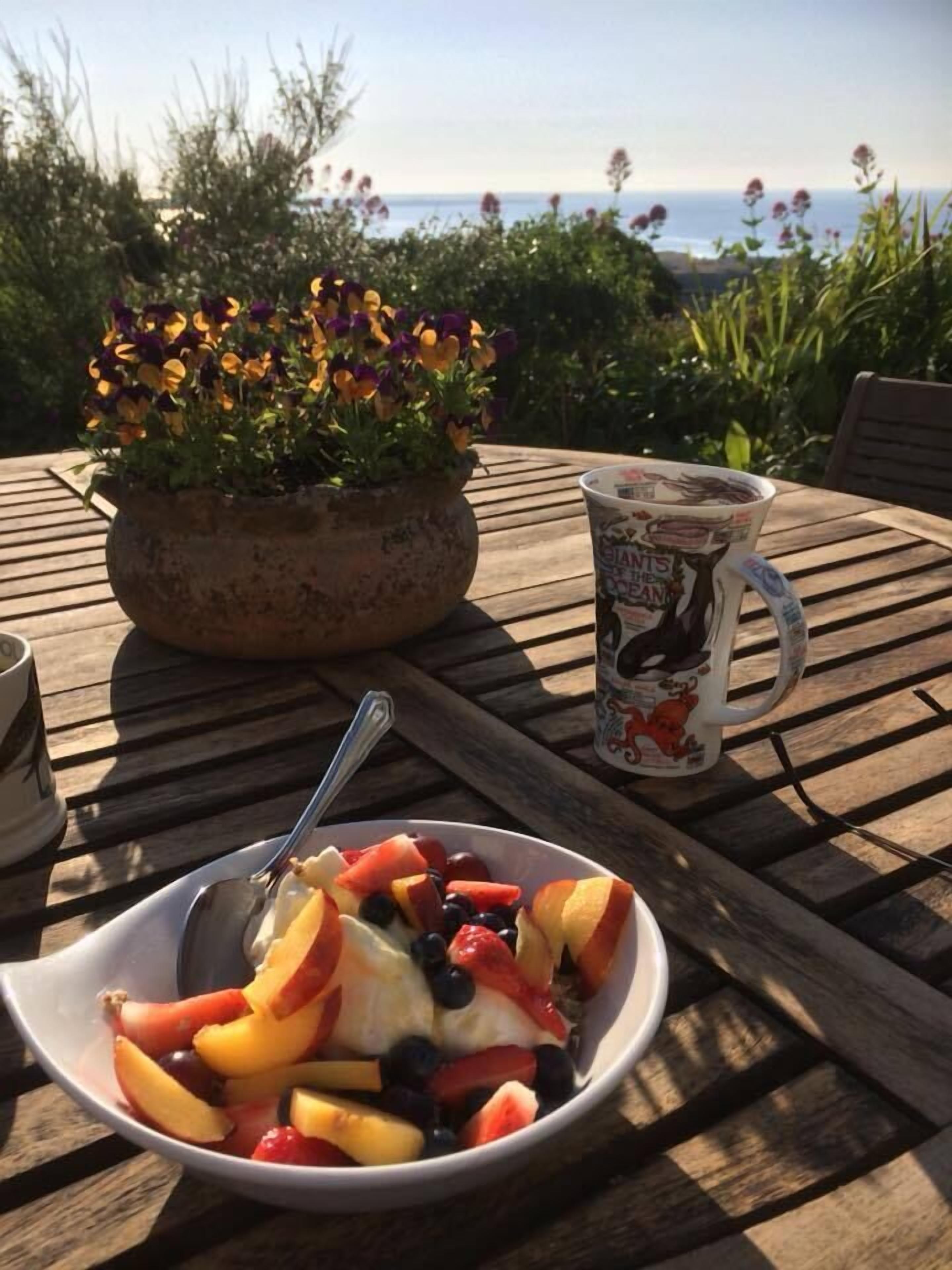A table with a bowl of fruit salad, a coffee mug, and a potted plant with flowers.