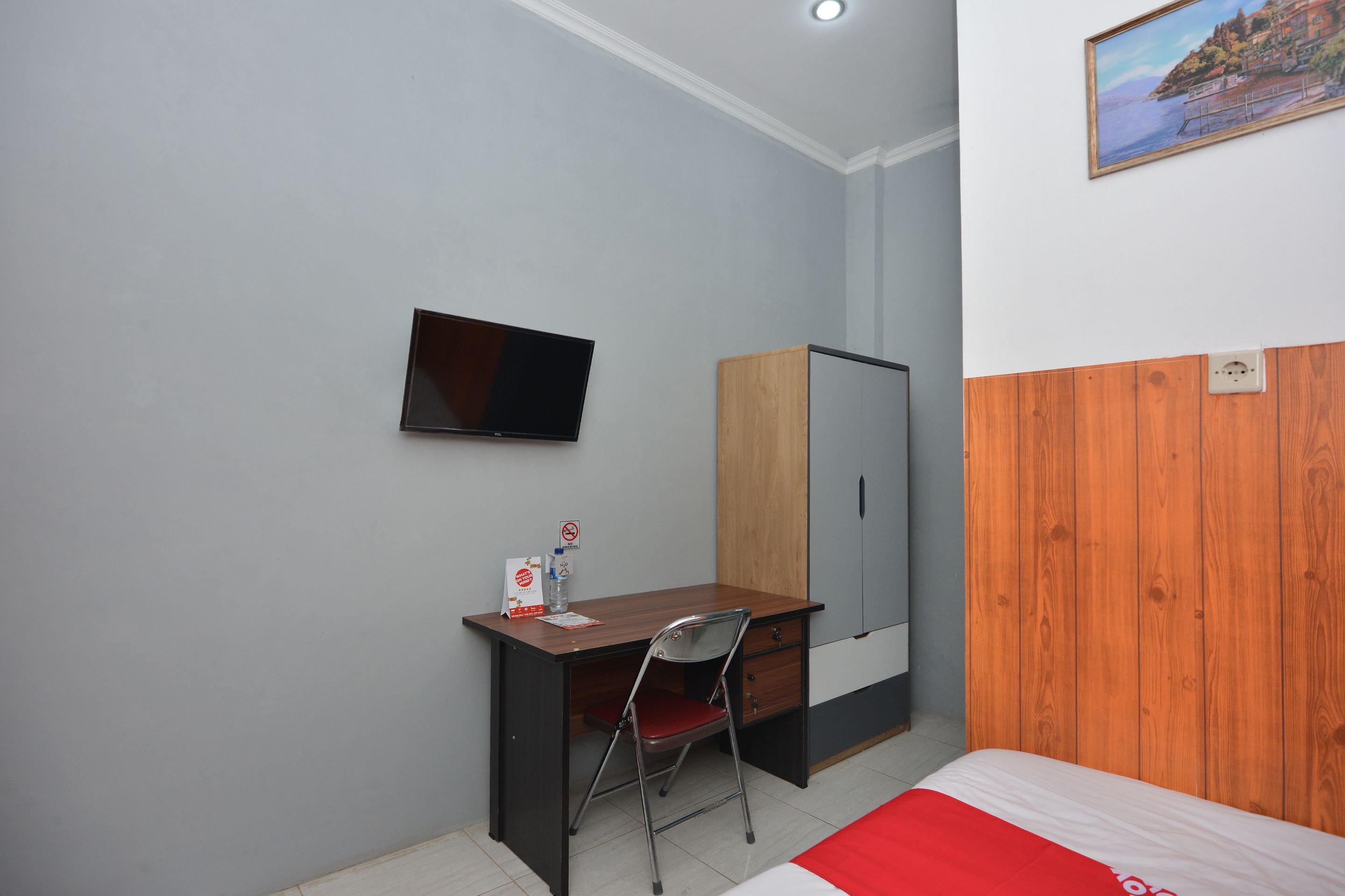 A hotel room with a wooden desk, a chair, a television mounted on the wall, a wardrobe, and a bed with a red blanket.