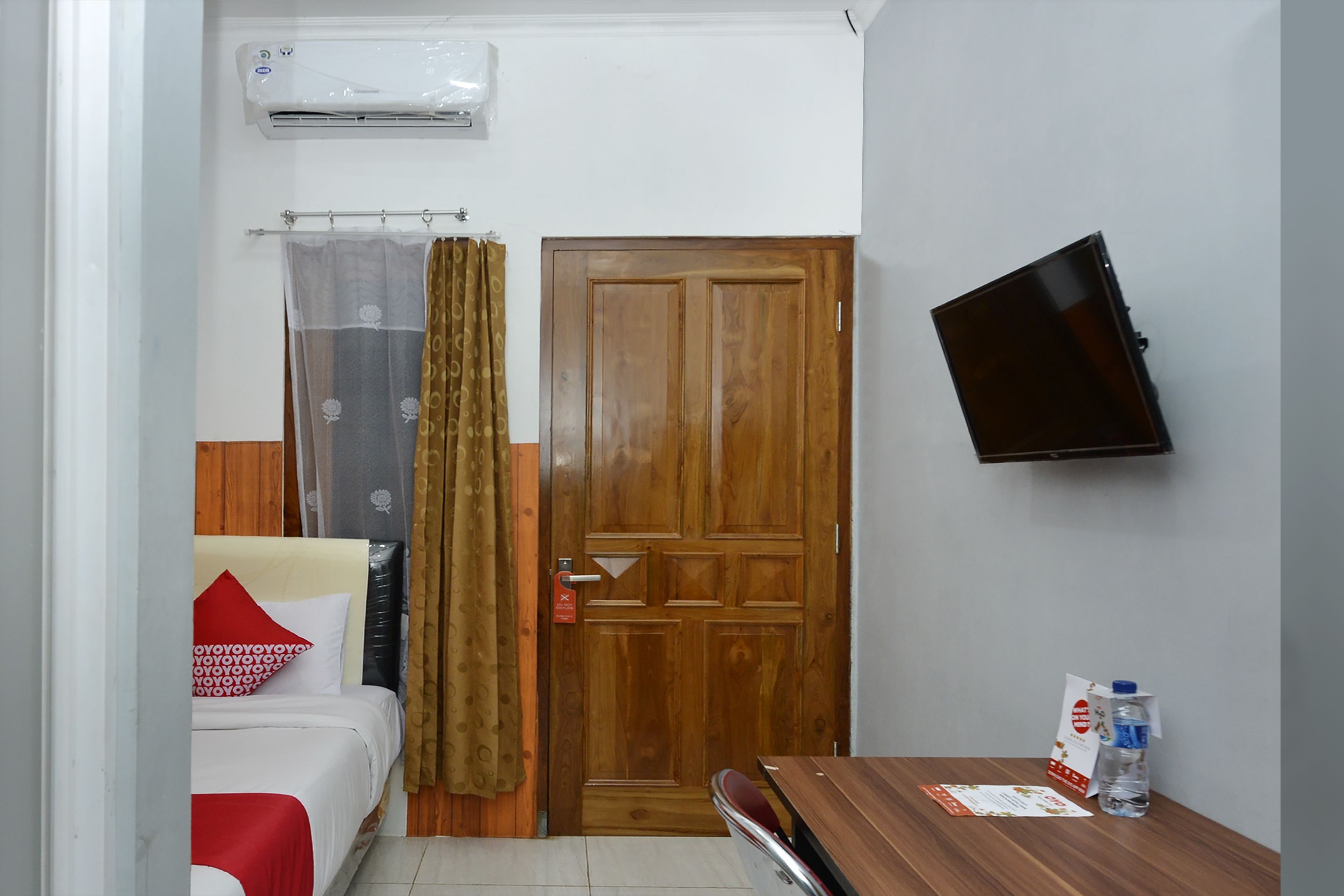Quarto de hotel pequeno com cama, porta de madeira, televisão na parede e mesa com uma garrafa e um menu.