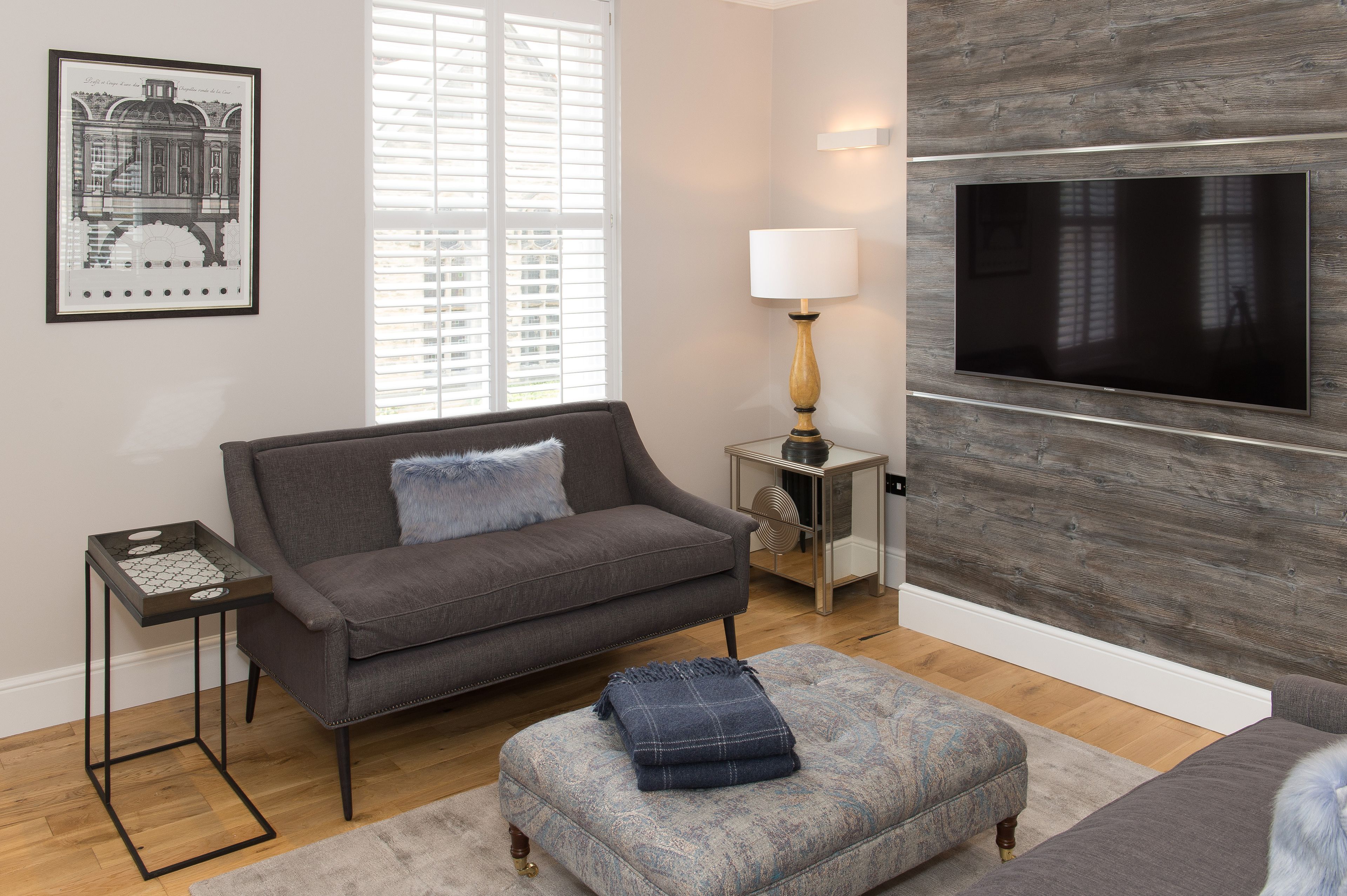 A modern living room with a grey sofa, a matching ottoman, a side table with a lamp, and a mounted TV.