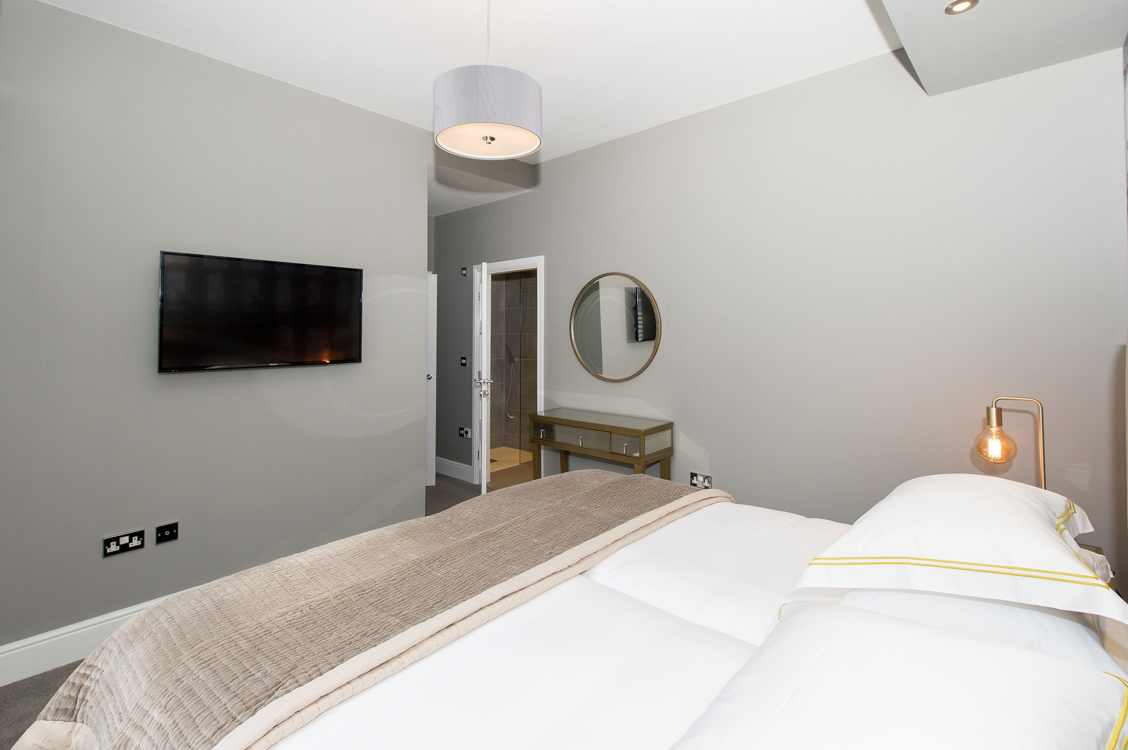 A modern hotel room with a bed, a TV mounted on the wall, a bedside table, a mirror, and a lamp.