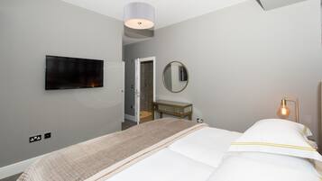 A modern hotel room with a bed, a TV mounted on the wall, a bedside table, a mirror, and a lamp.