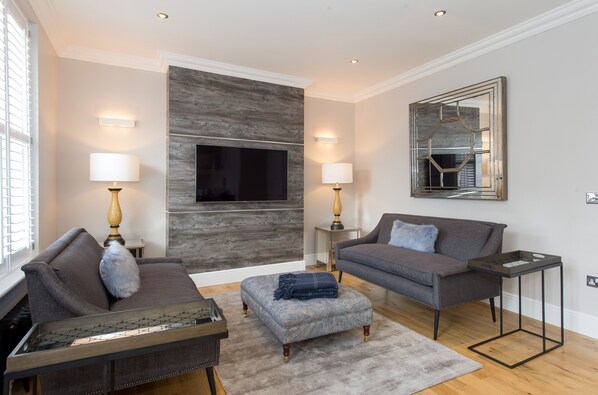 A modern living room with a grey sofa, a matching ottoman, a coffee table, and a TV mounted on a wooden wall. There are two floor lamps and a side table with a lamp. - Belford (Harrogate)