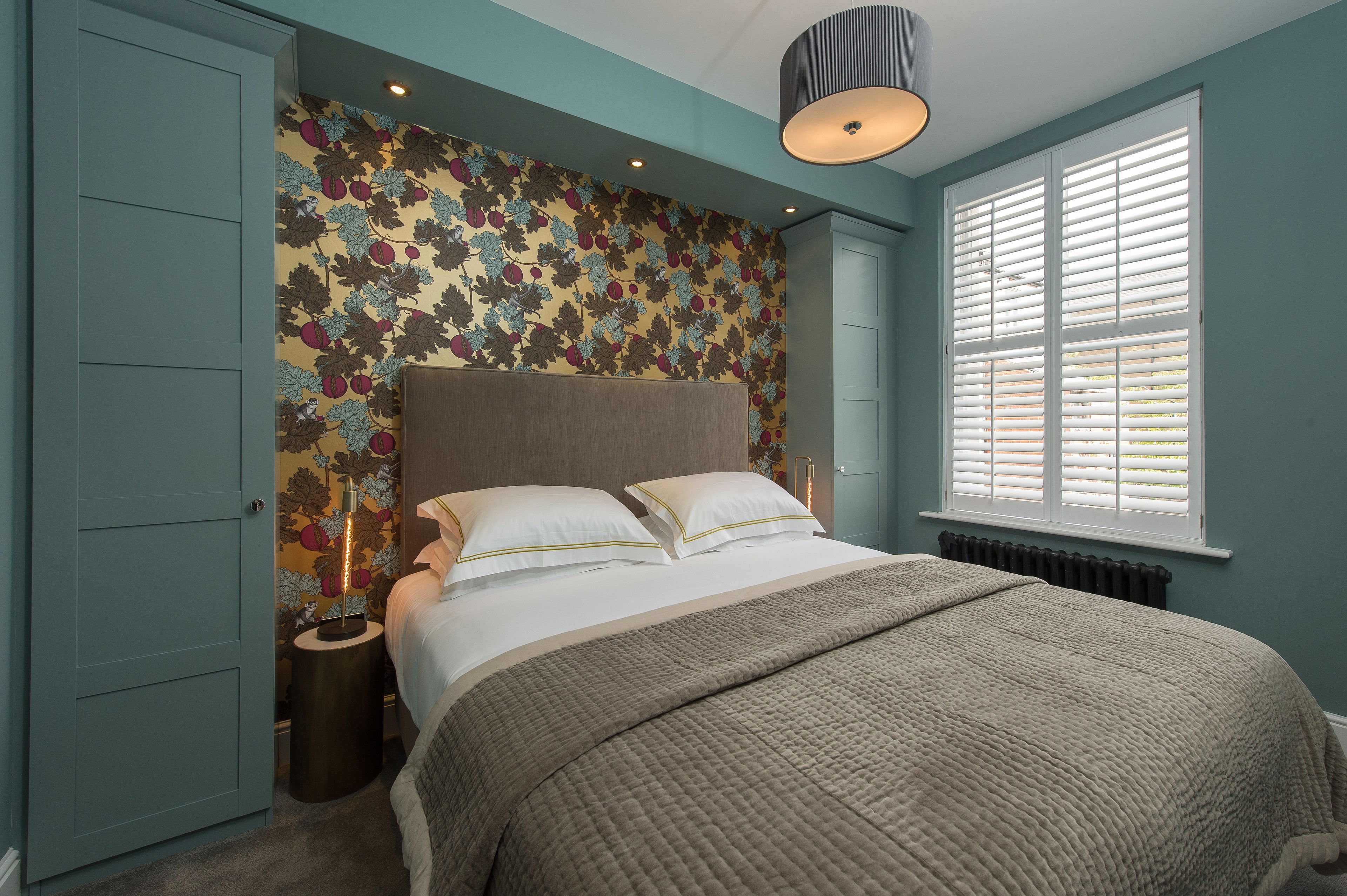 A bedroom with a bed, a nightstand, a wall with a floral pattern, a window with blinds, and a ceiling light.
