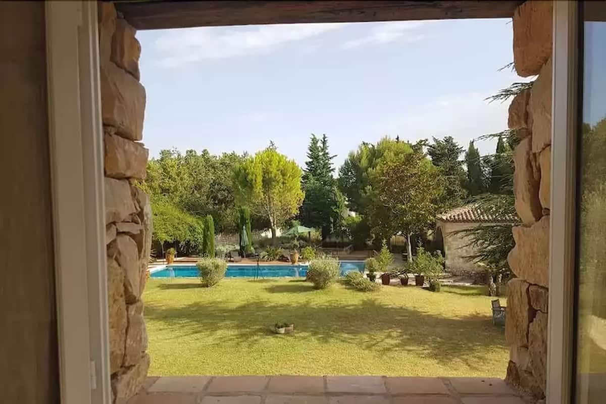 Vista a través de una ventana enmarcada en piedra de un jardín trasero con piscina, césped verde y varios árboles.