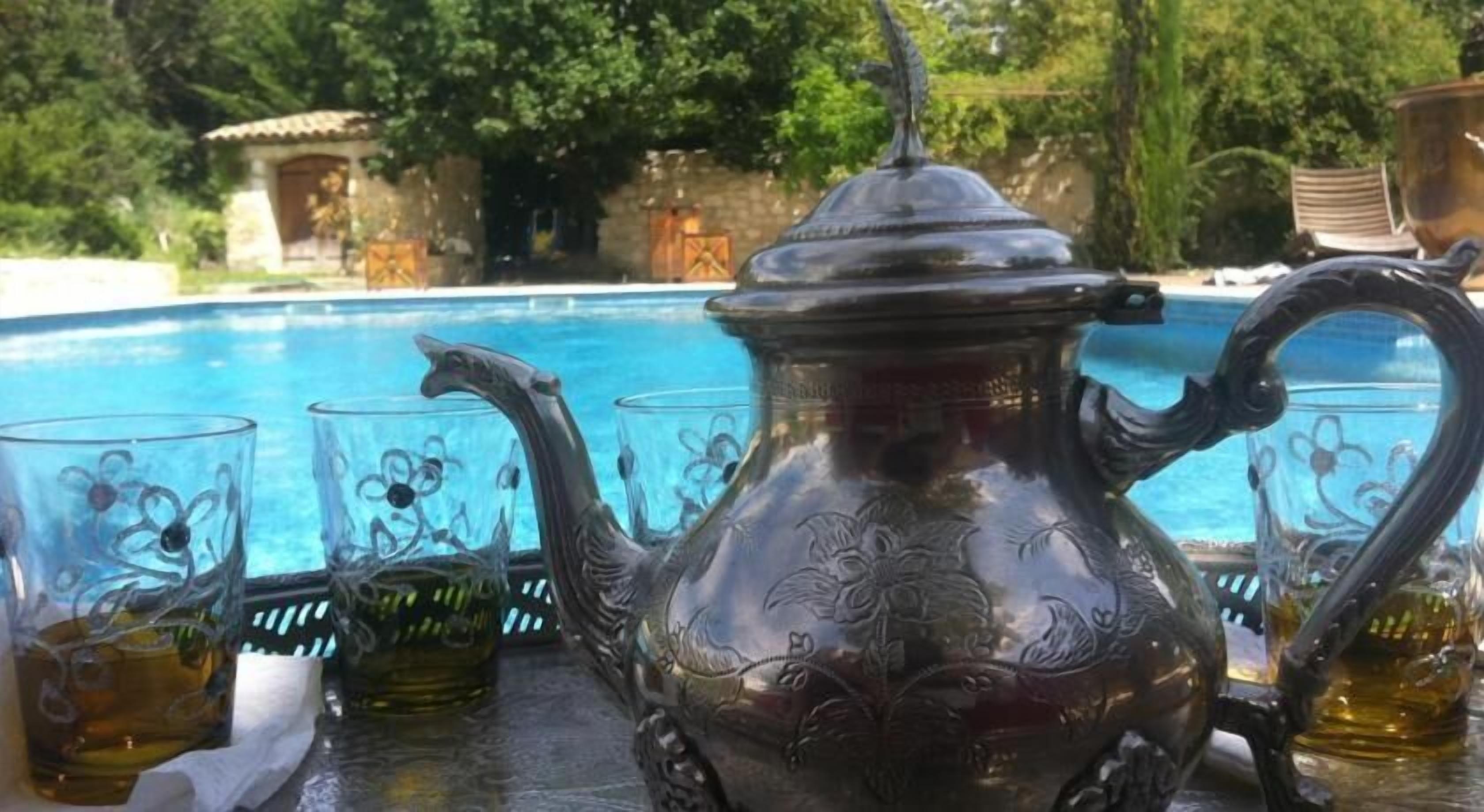 Une théière en métal aux motifs complexes, accompagnée de quatre verres aux motifs floraux, est posée sur une table près d’une piscine.