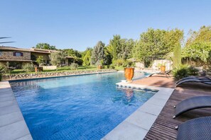Une piscine dotée d’une terrasse en bois et de chaises longues, entourée de verdure et bordée d’un bâtiment en pierre.