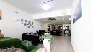A reception area with a white wall, framed pictures, a large white vase with flowers, a green plant, and a black sofa.