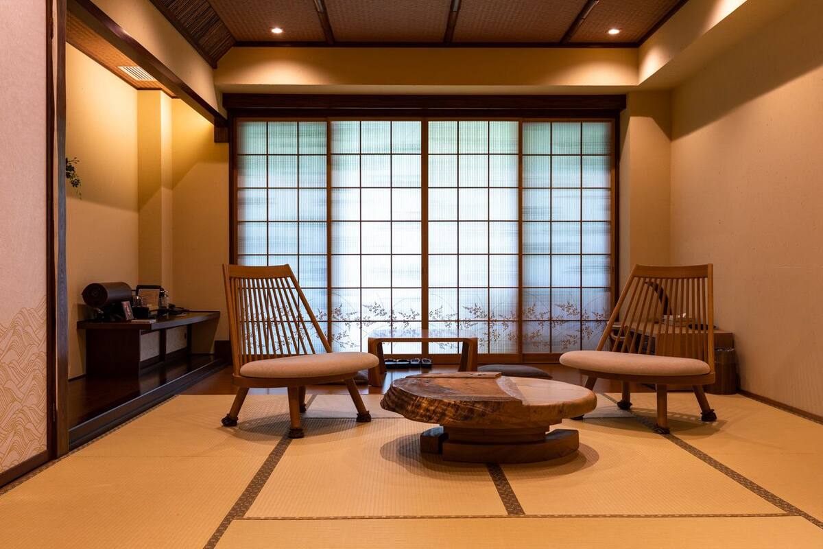 A traditional Japanese room with tatami flooring, two wooden chairs, a low wooden table, and a shoji screen window.