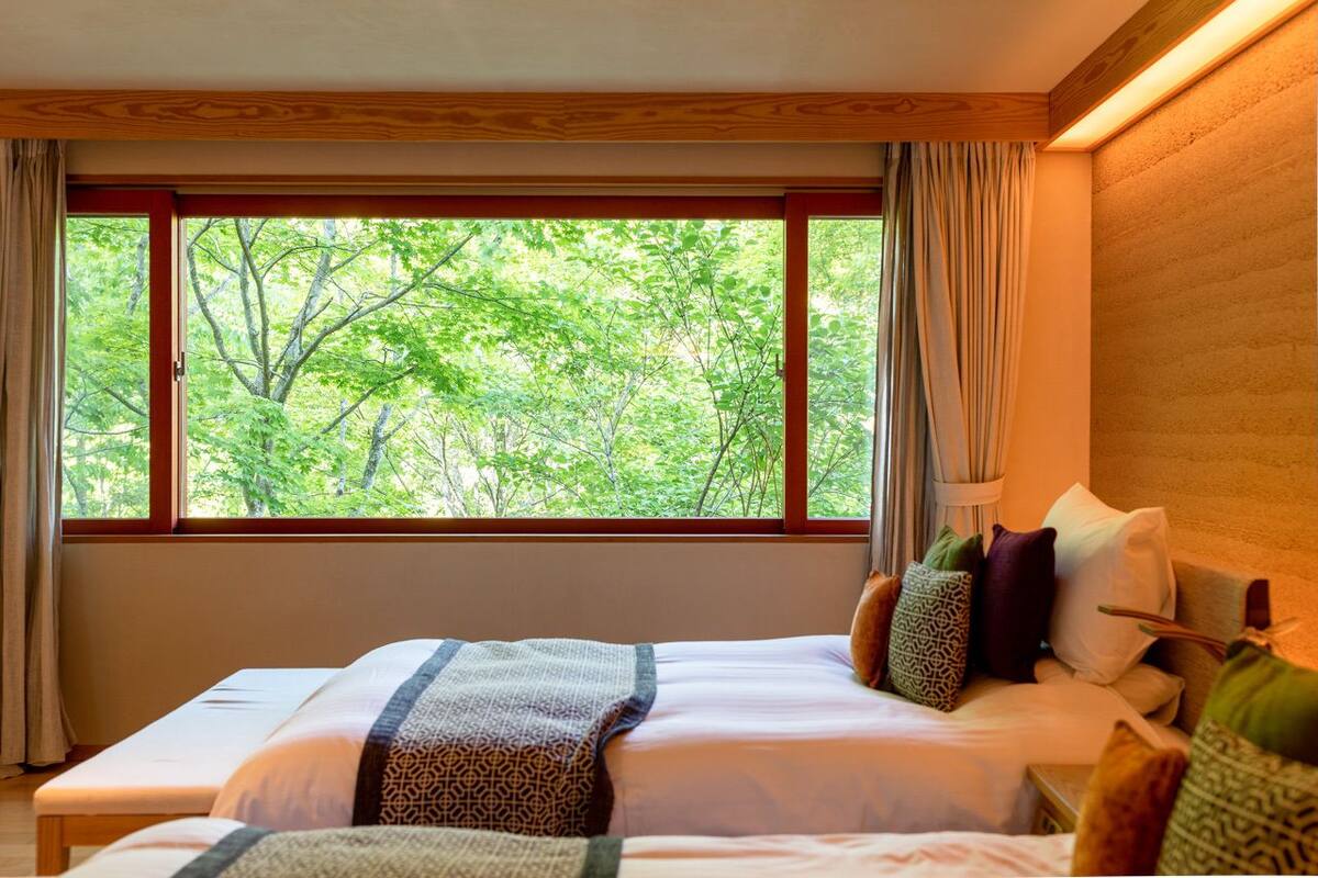 A hotel room with a large window offering a view of trees. The room has a bed with patterned bedding and several pillows.