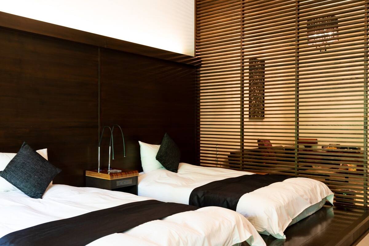A modern hotel room with two beds, wooden headboard, and a large window with blinds.