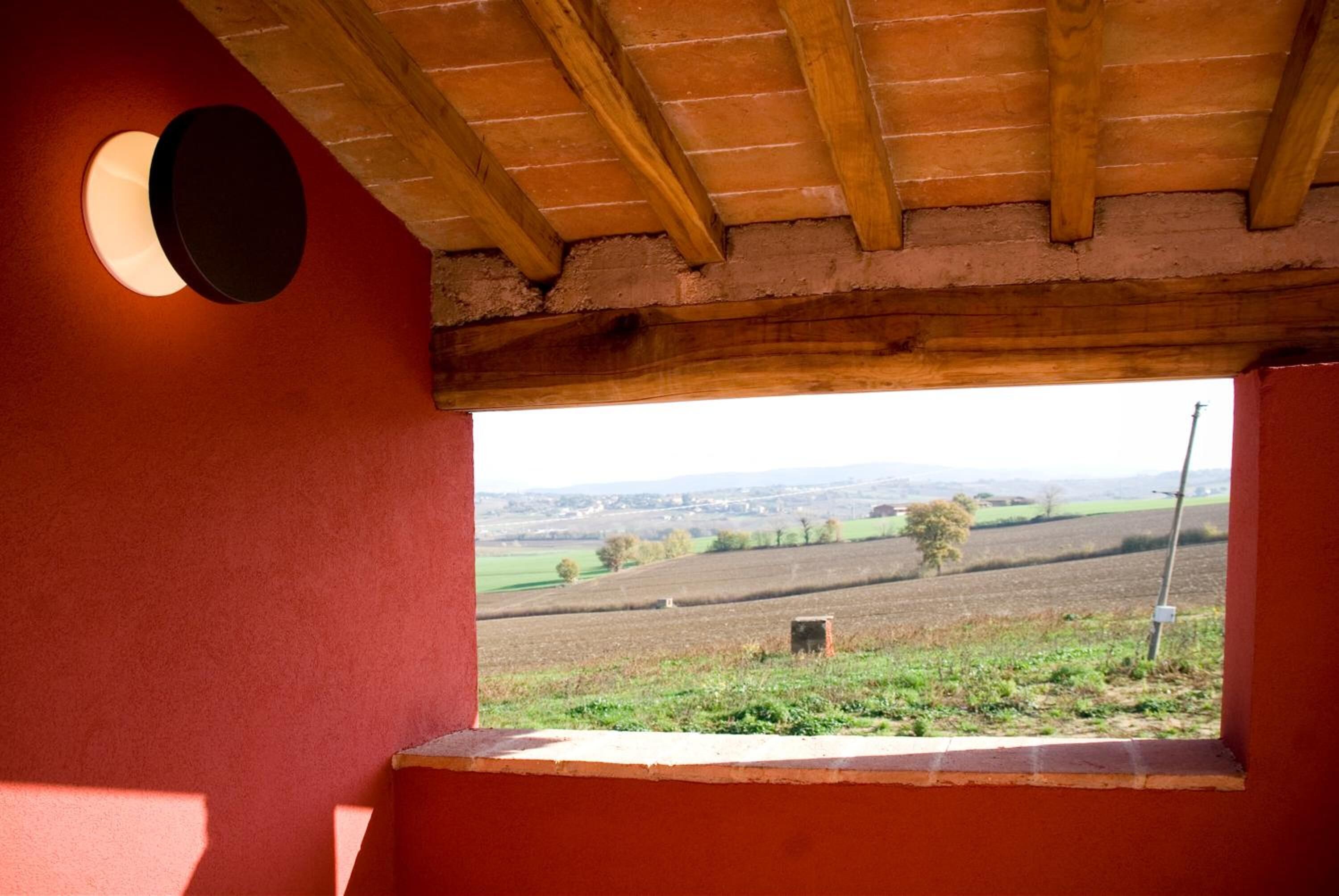 Een kamer met een raam dat uitkijkt op een landelijk landschap, houten balken in het plafond en een rode muur.