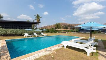 A swimming pool with lounge chairs and umbrellas, surrounded by a landscaped area with palm trees and a building in the background.