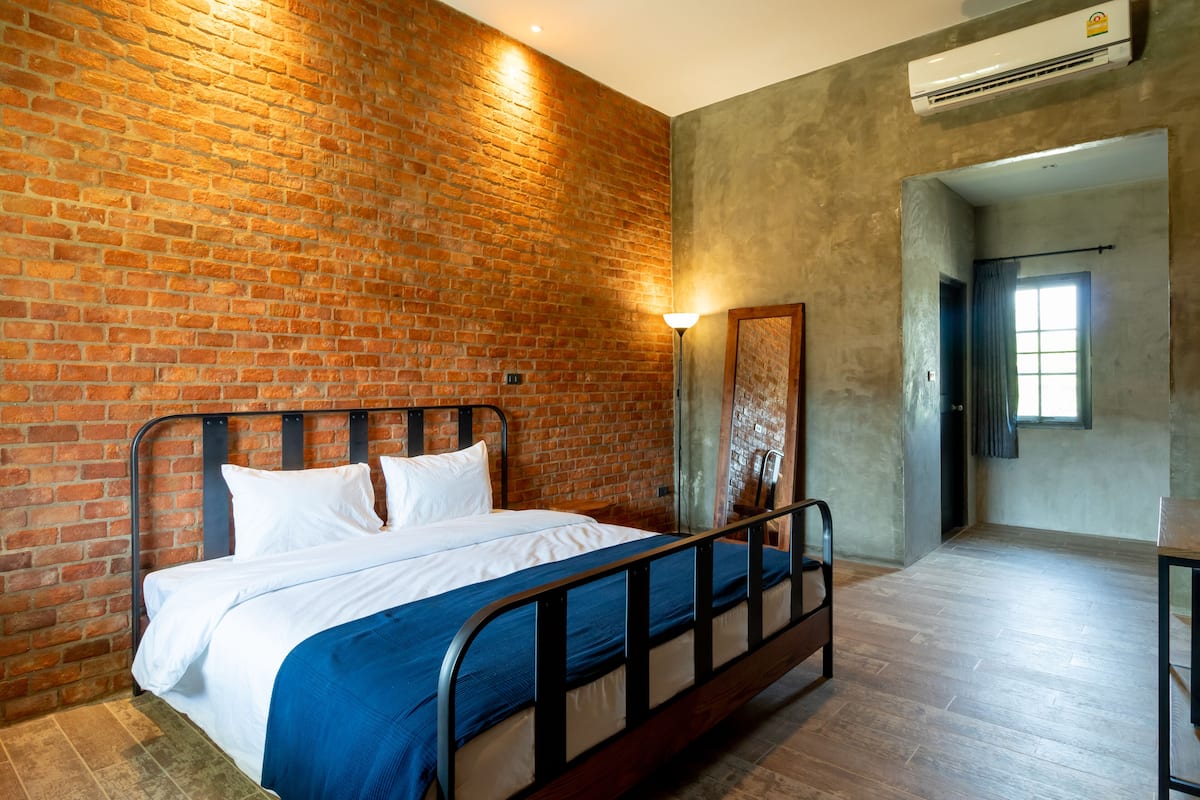 Quarto com parede de tijolos, cama com roupa de cama branca, manta azul, cabeceira de metal e chão de madeira, além de um aparelho de ar condicionado.