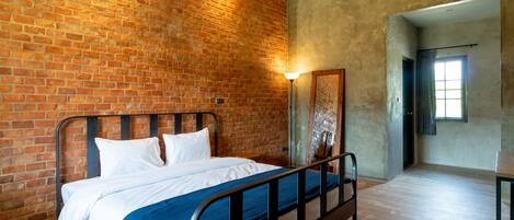 Una habitación con pared de ladrillo, cama con ropa de cama blanca, manta azul, cabecero de metal, piso de madera y un equipo de aire acondicionado.