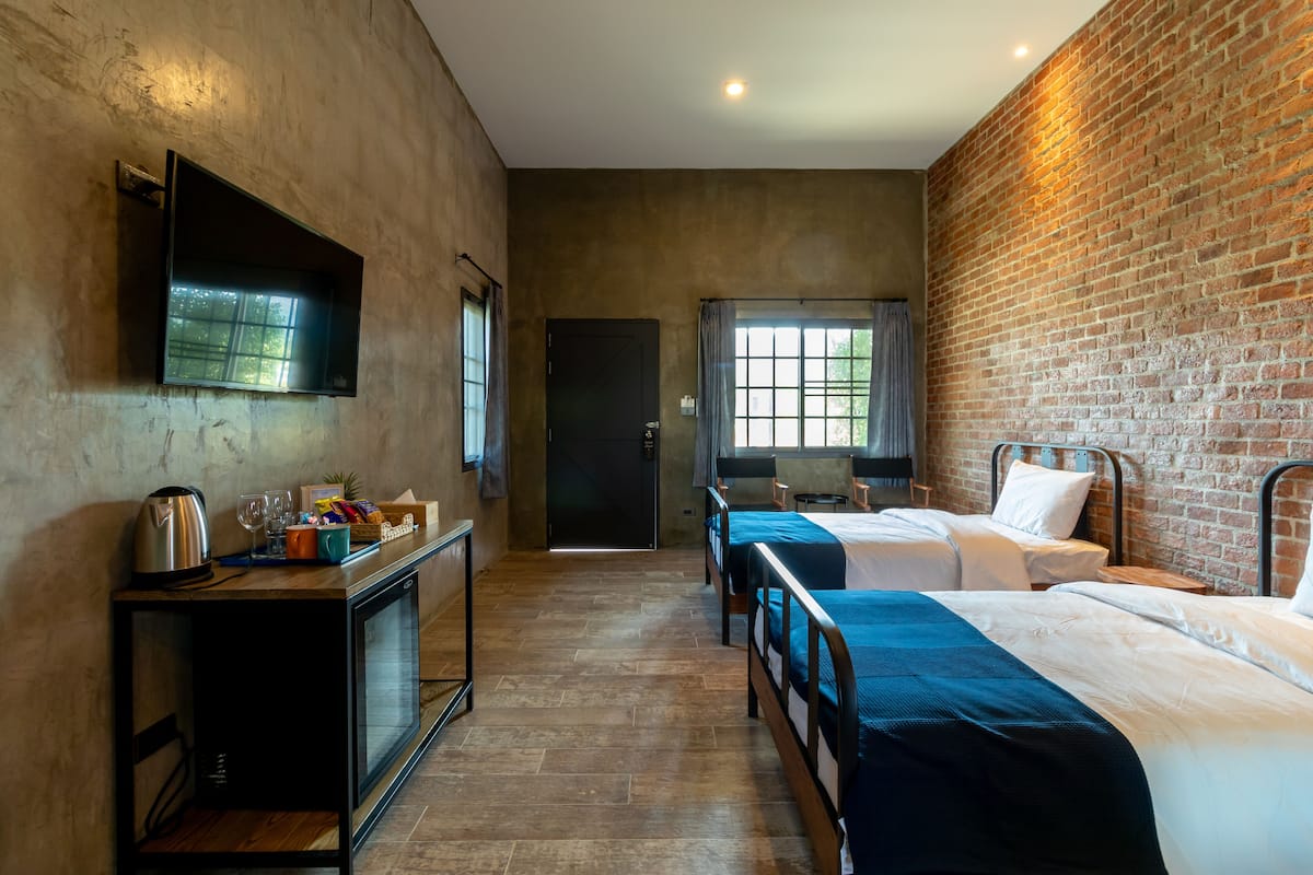 Quarto de hotel com duas camas, televisão, pequeno espaço com cozinha e parede de tijolos.
