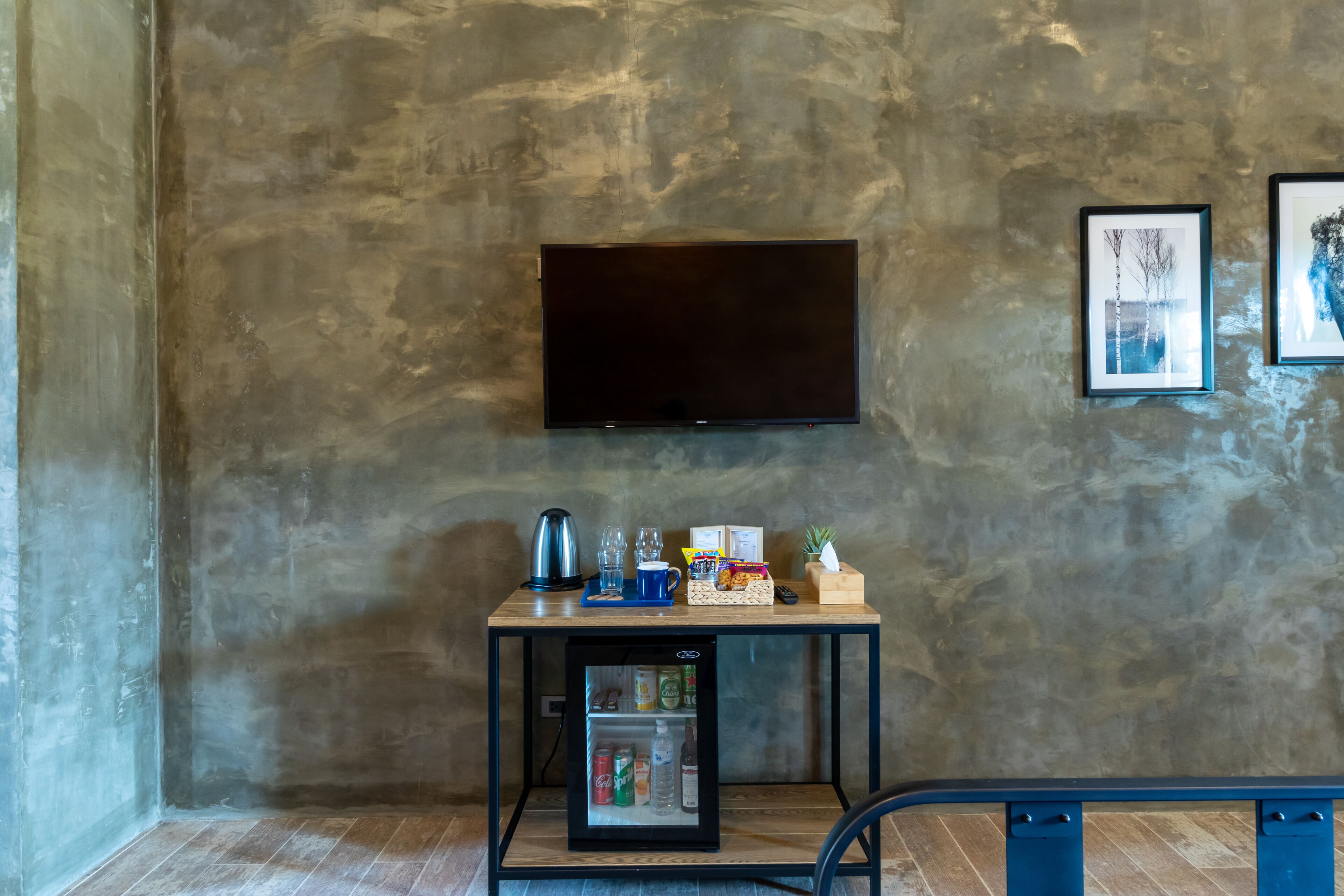 A compact room with a flat-screen TV mounted on the wall, a small wooden table with a mini-fridge, a kettle, and some snacks.