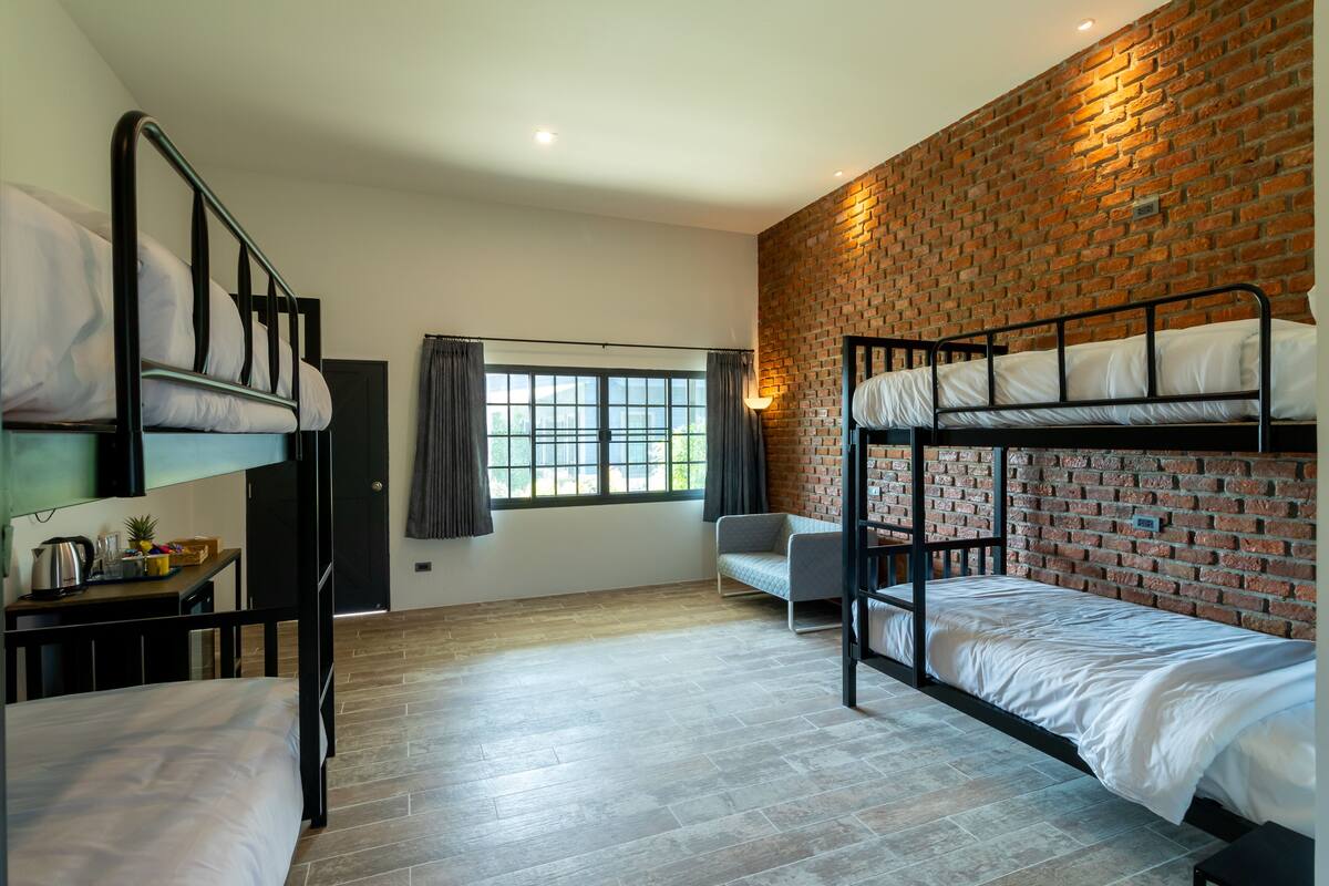 Um quarto em dormitório com beliches, uma escrivaninha com chaleira, uma cadeira e uma janela com cortinas.
