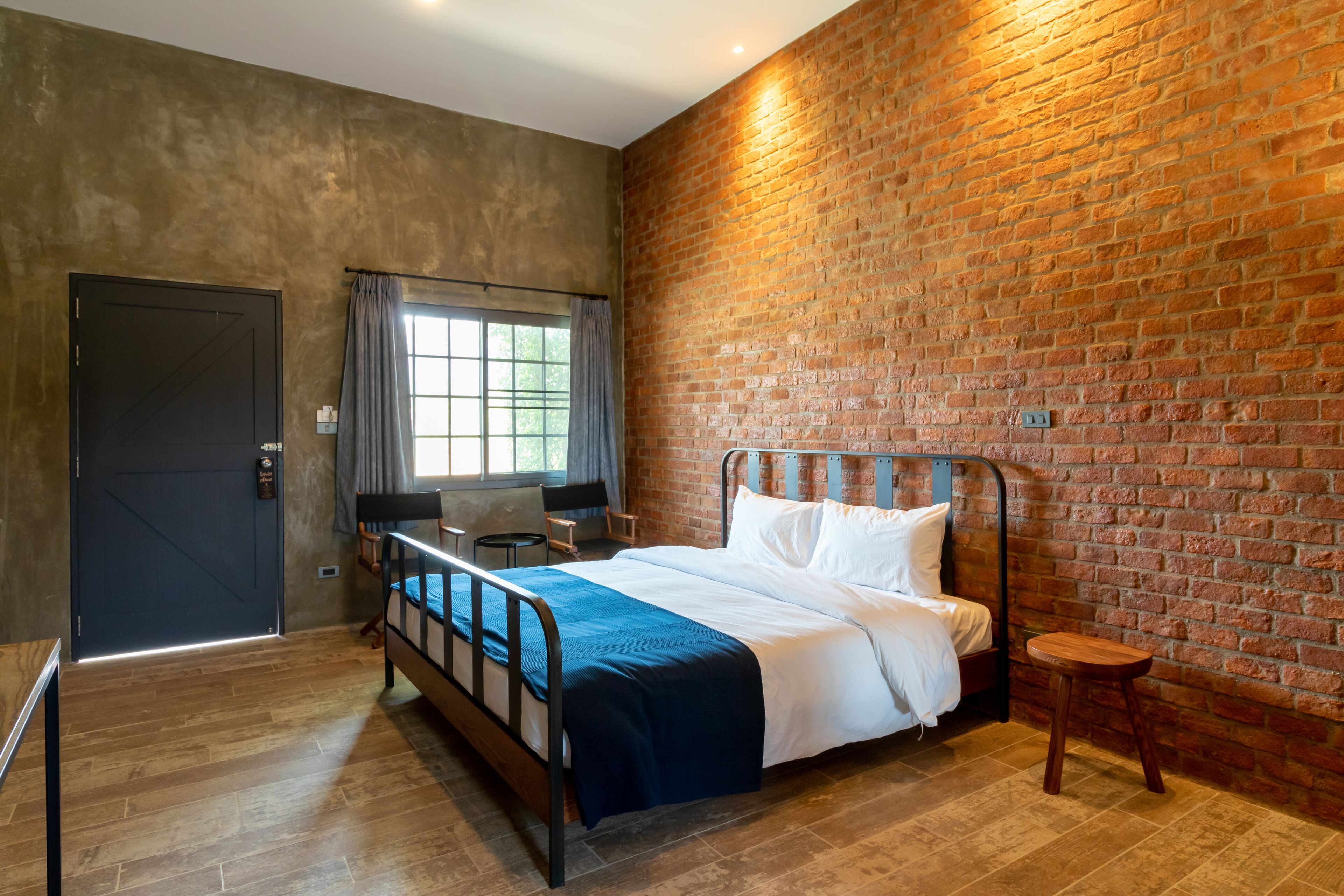 Un dormitorio con pared de ladrillo, cama con sábanas blancas, piso de madera, ventana con cortinas y una puerta.