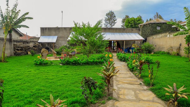 Un camino de piedra que lleva a una casa con techo de metal, rodeada por un jardín verde exuberante con diversas plantas y una pequeña construcción con techo de tejas.