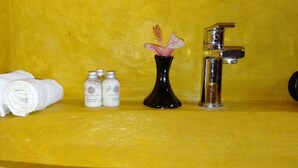 Un lavamanos con una pared amarilla, un florero negro con una flor rosa, dos botellas de loción corporal y una toalla blanca enrollada.