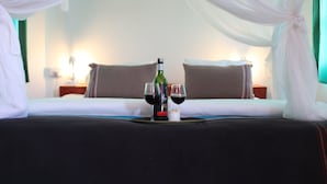 A hotel room with a bed, two pillows, a bedside table with a lamp, and a tray with a bottle and two glasses of red wine. - Dan Stam Hotel (Arusha)