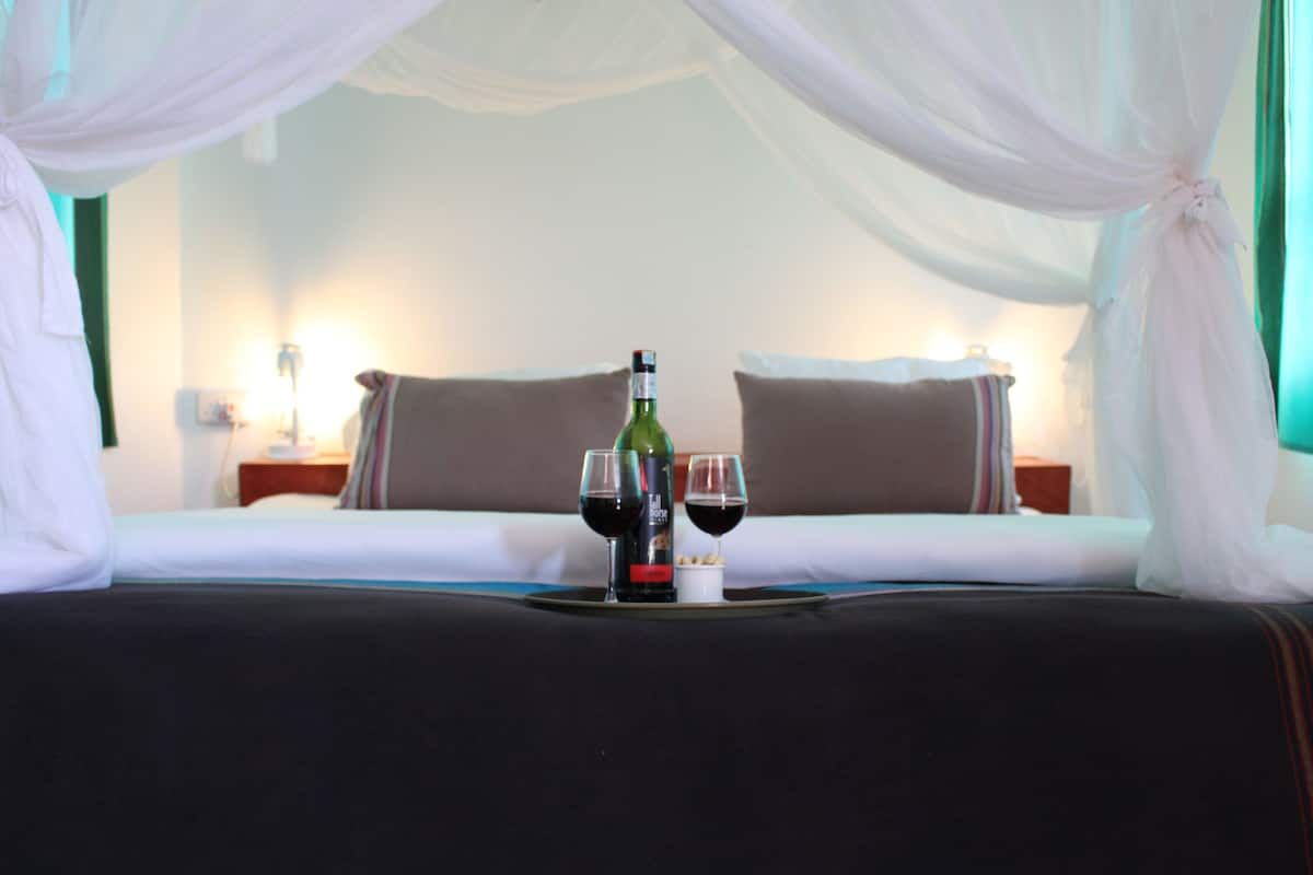 A hotel room with a bed, two pillows, a bedside table with a lamp, and a tray with a bottle and two glasses of red wine.