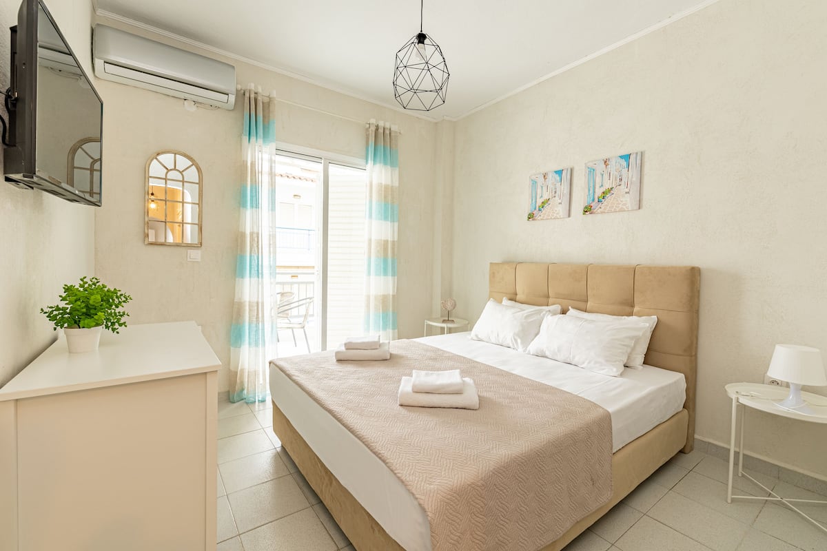 A modern bedroom with a large bed, a bedside table, a small table, a potted plant, and a window with curtains.