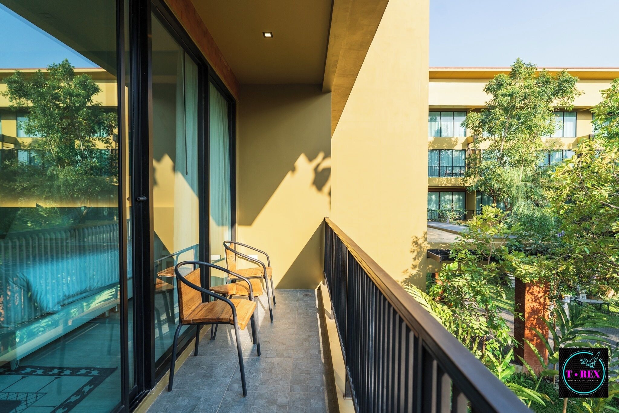 A balcony with two wooden chairs and a glass door, overlooking a courtyard with trees and a building in the background.