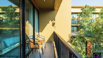A balcony with two wooden chairs and a glass door, overlooking a courtyard with trees and a building in the background.