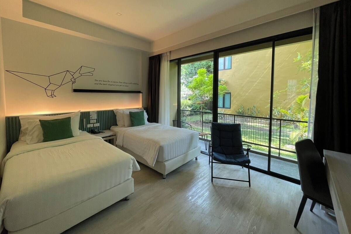 Habitación de hotel con dos camas, una silla y balcón con vistas a la vegetación.