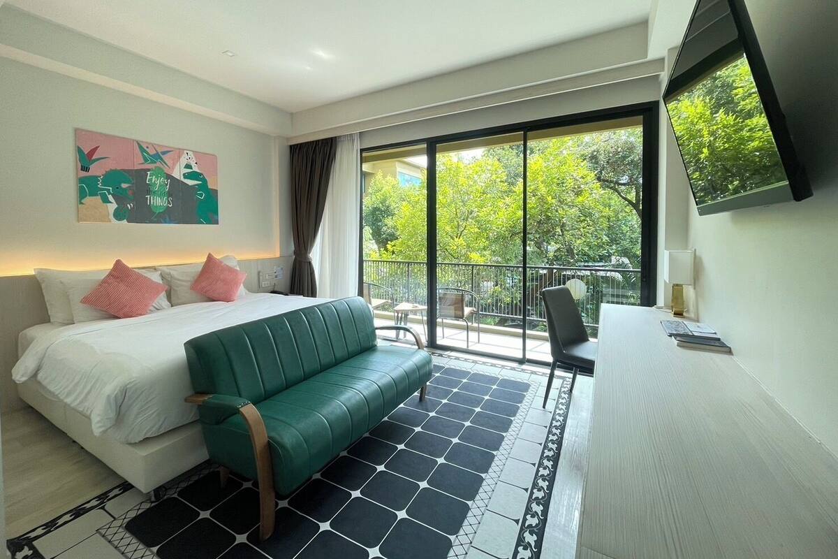 Una habitación de hotel moderna con cama, sofá verde, escritorio con silla y ventana grande con vistas a árboles.
