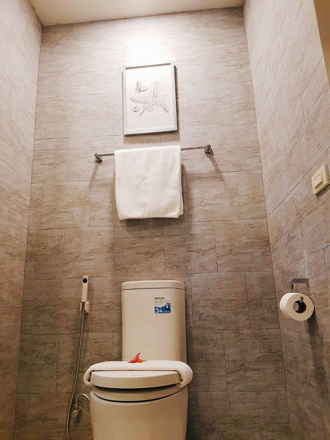 Un baño con inodoro, un cuadro en la pared, una toalla colgada en un perchero y un dispensador de papel higiénico.