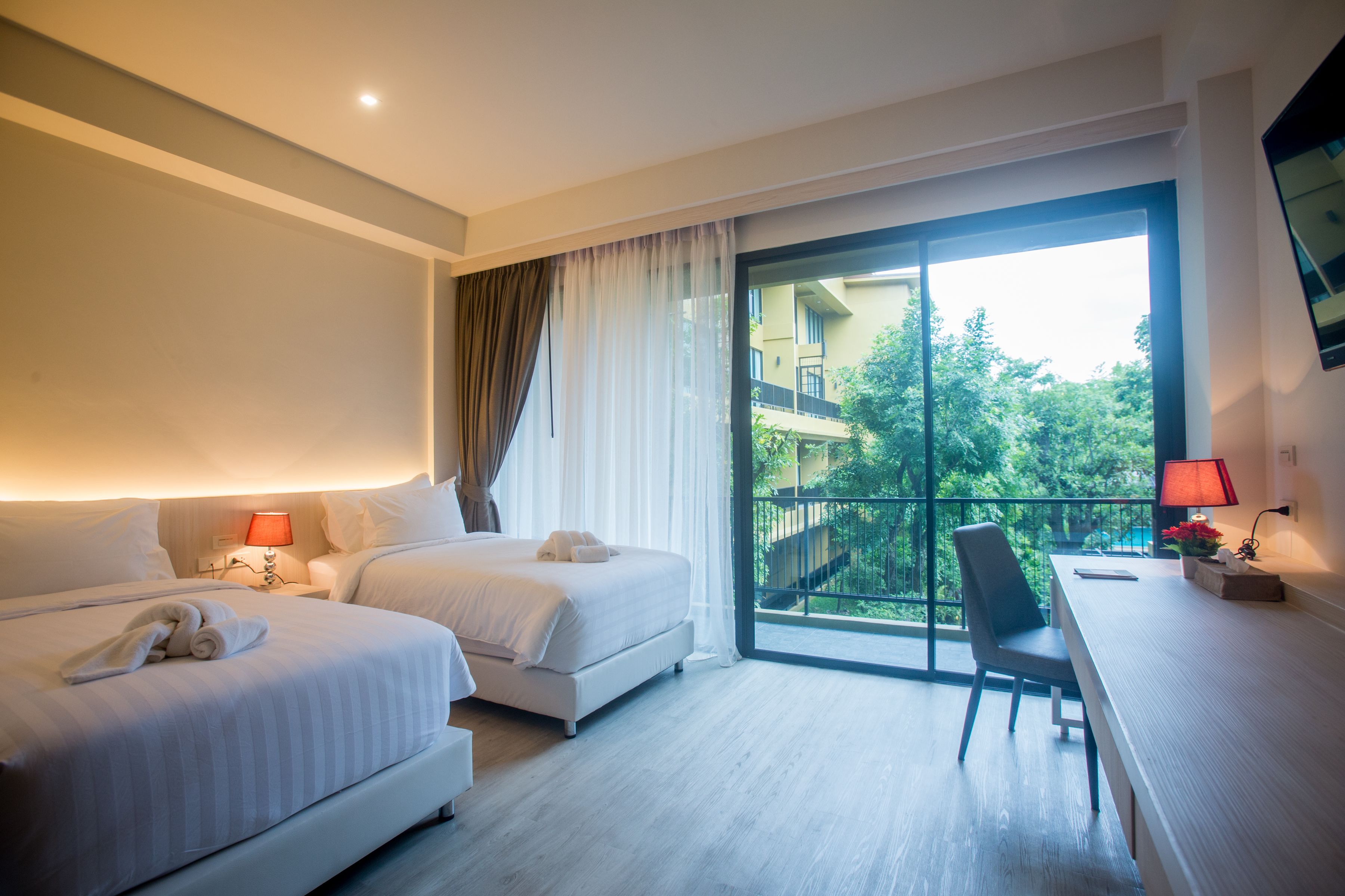 A hotel room with two beds, a desk, a chair, a balcony with a view of greenery, and a TV mounted on the wall.
