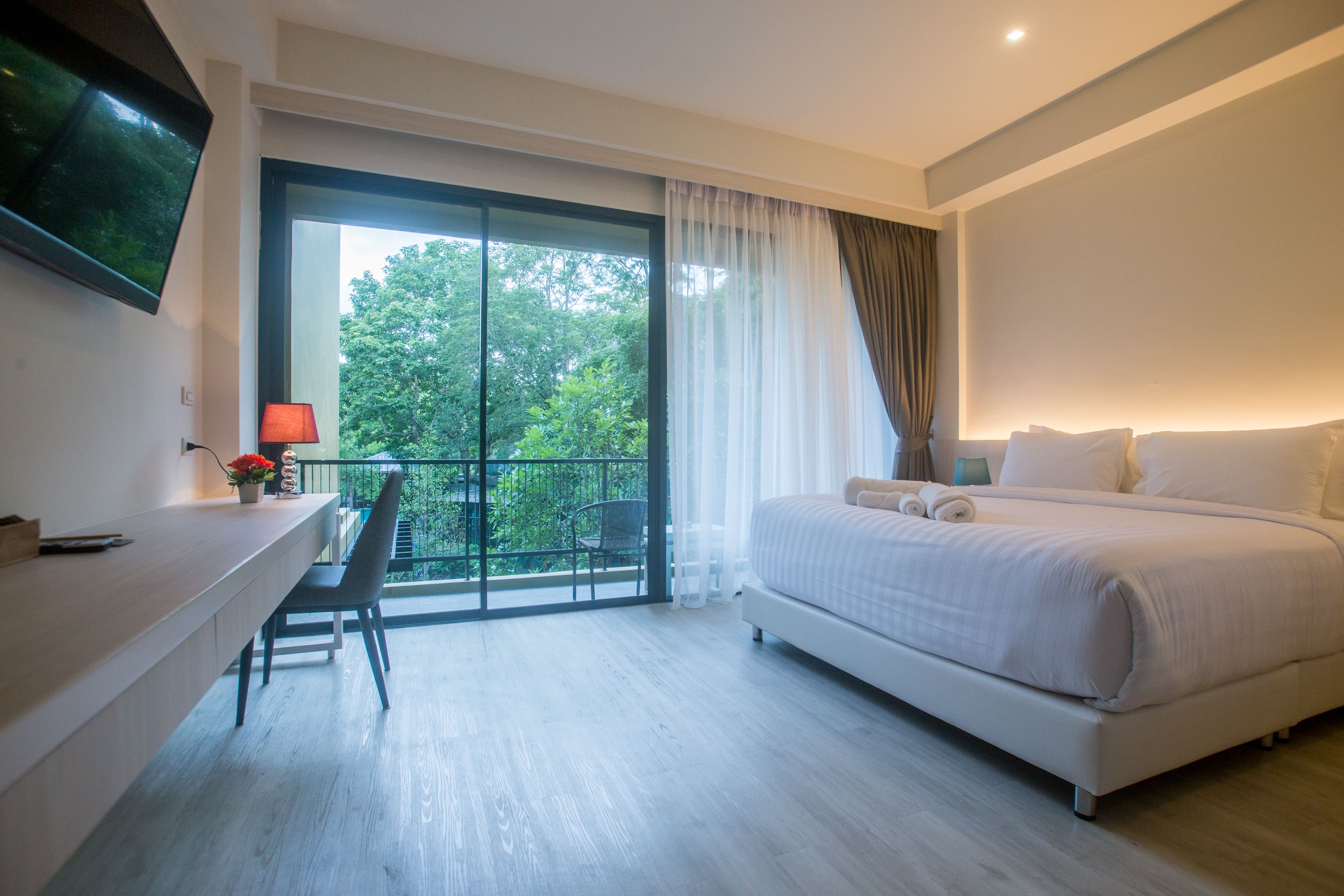 A modern hotel room with a large bed, a desk with a chair, a TV mounted on the wall, and a balcony with a view of trees.
