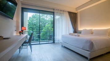 A modern hotel room with a large bed, a desk with a chair, a TV mounted on the wall, and a balcony with a view of trees.