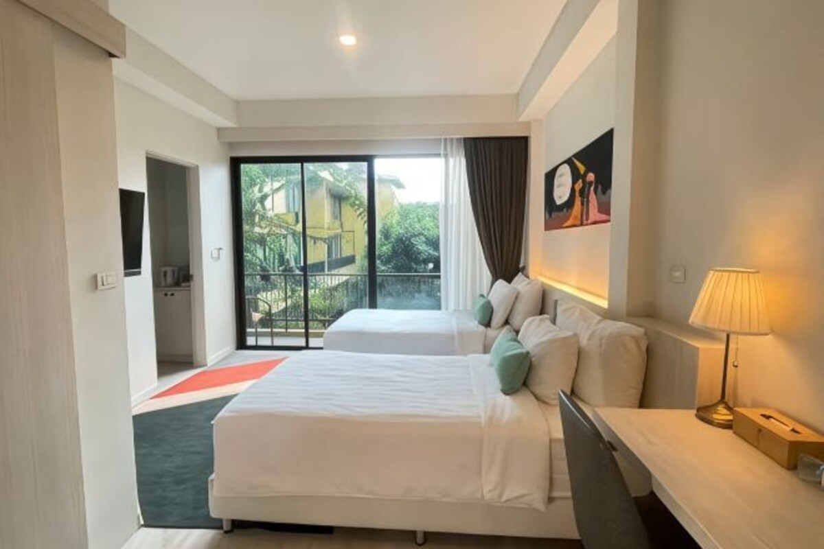 A hotel room with two beds, a desk, a lamp, and a view of greenery outside.