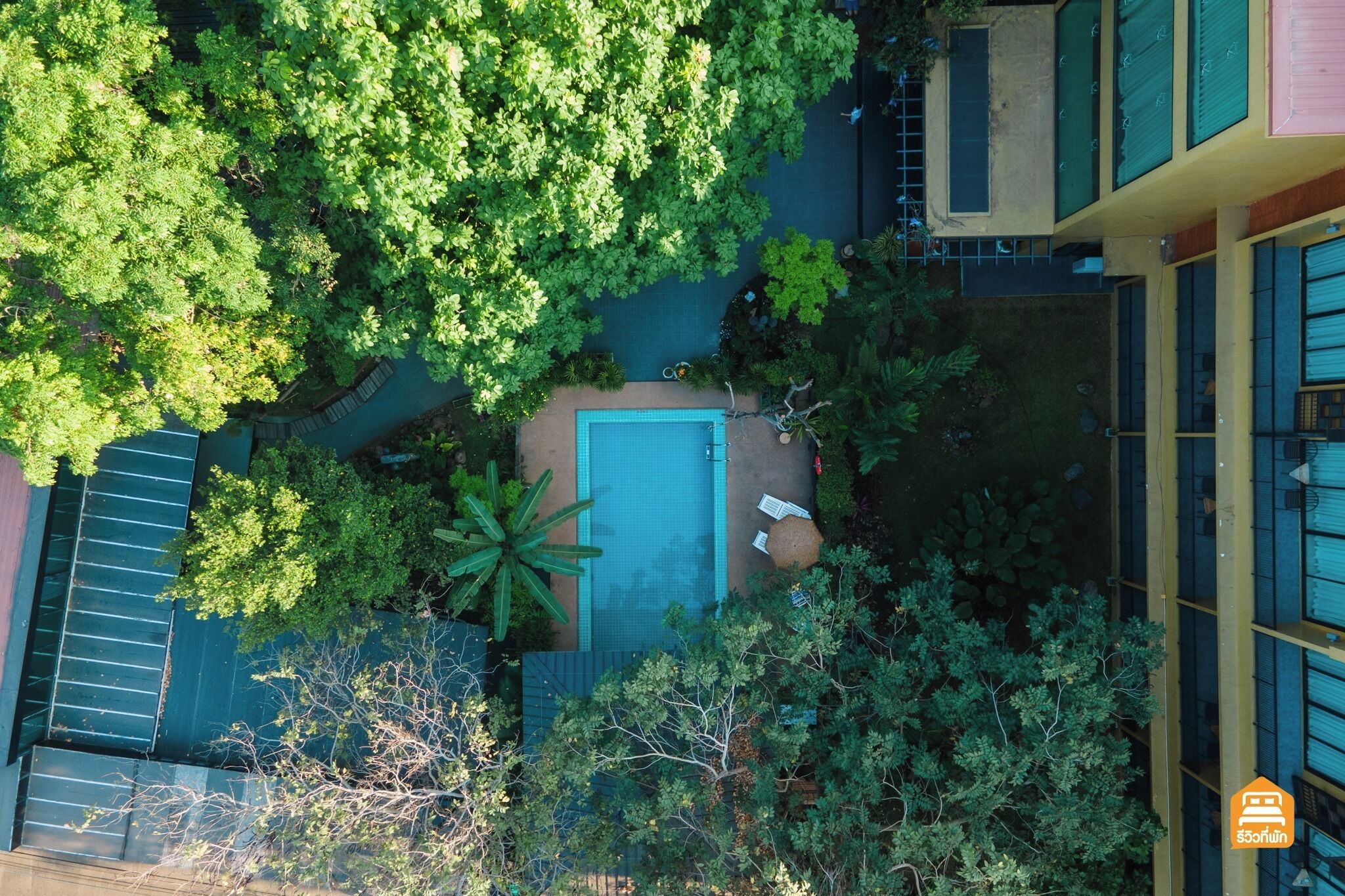 Vista aérea de un hotel con piscina rodeada de exuberante vegetación, un porche cubierto y un edificio de varios pisos con grandes ventanales.