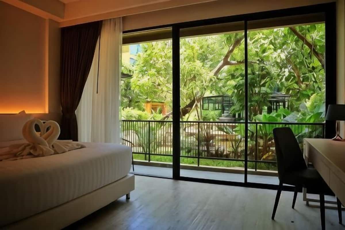 Habitación de hotel con una cama, un escritorio y vistas a un jardín a través de ventanas amplias.