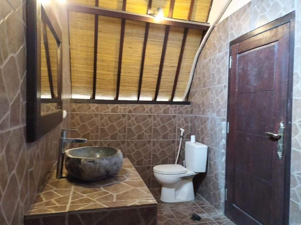 A bathroom with a stone wall, a wooden ceiling, a large mirror, a stone sink, a toilet, and a dark wooden door.
