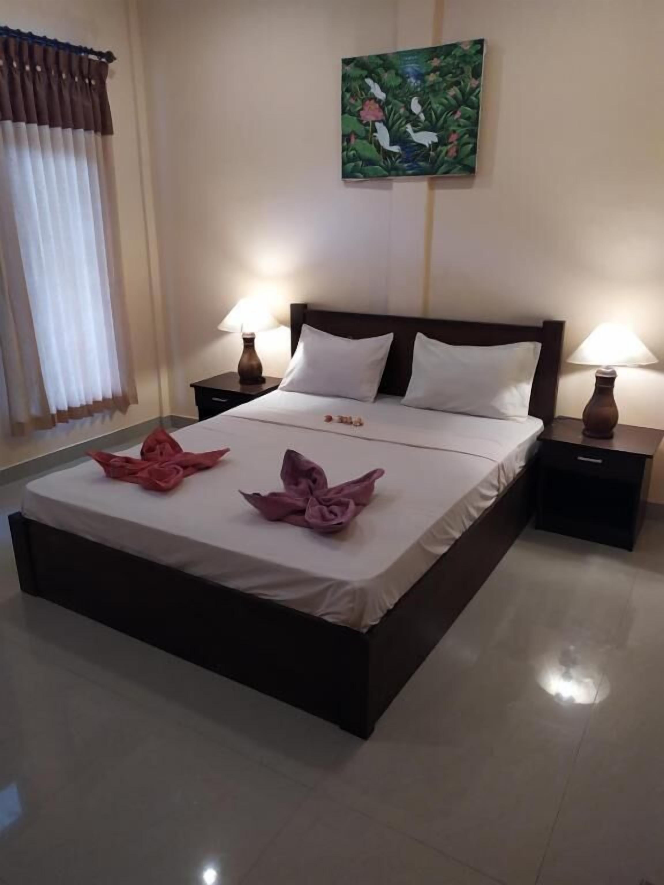 A neatly made bed with white linens, two pillows, and pink towels arranged on the bed. There are two bedside tables with lamps, a painting on the wall, and a window with curtains.
