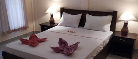 A neatly made bed with white linens, two pillows, and pink towels arranged on the bed. There are two bedside tables with lamps, a painting on the wall, and a window with curtains.