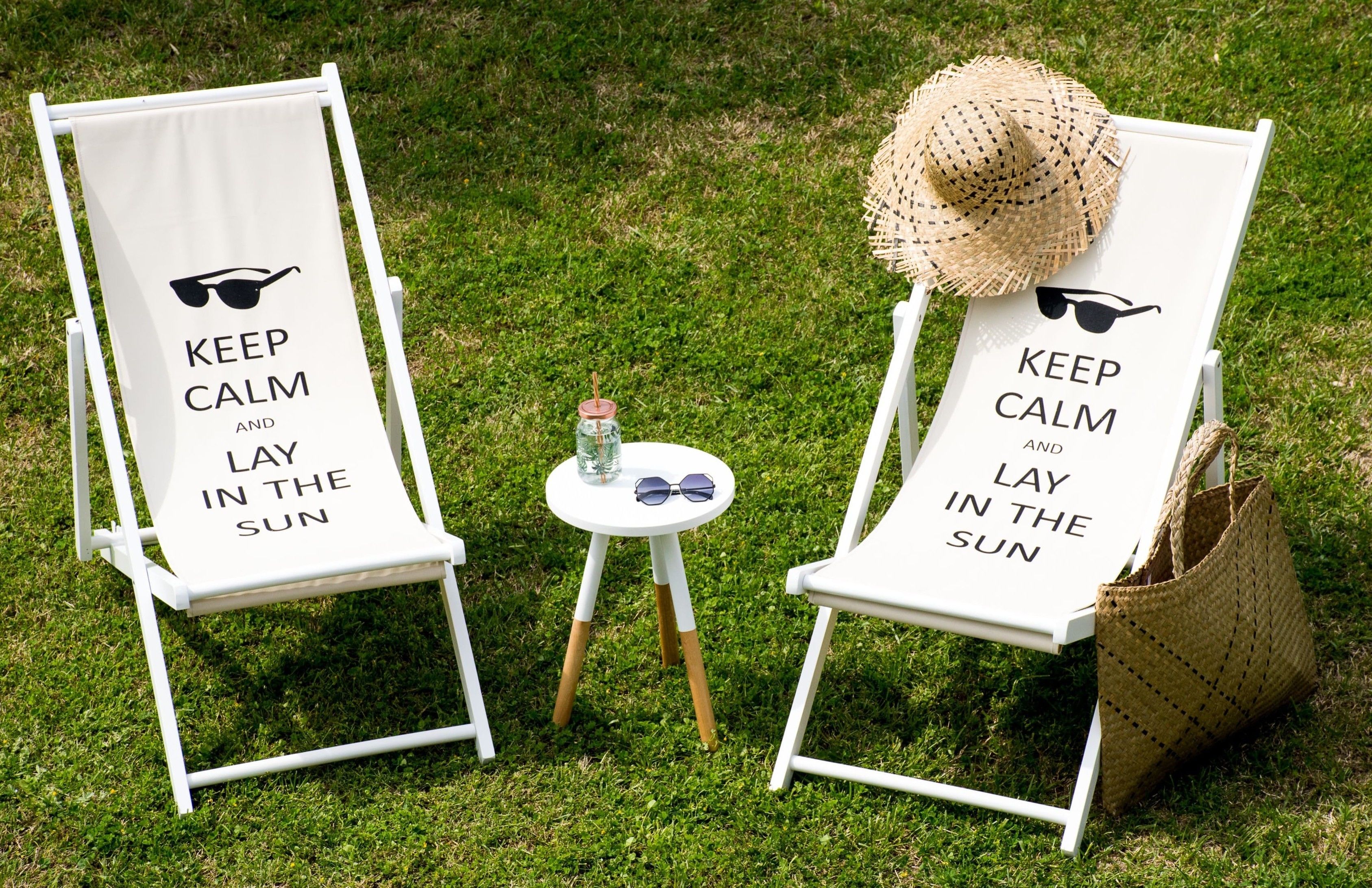 Dos sillones blancos con la inscripción "KEEP CALM AND LAY IN THE SUN", ubicados sobre un césped con una pequeña mesa entre ellos.