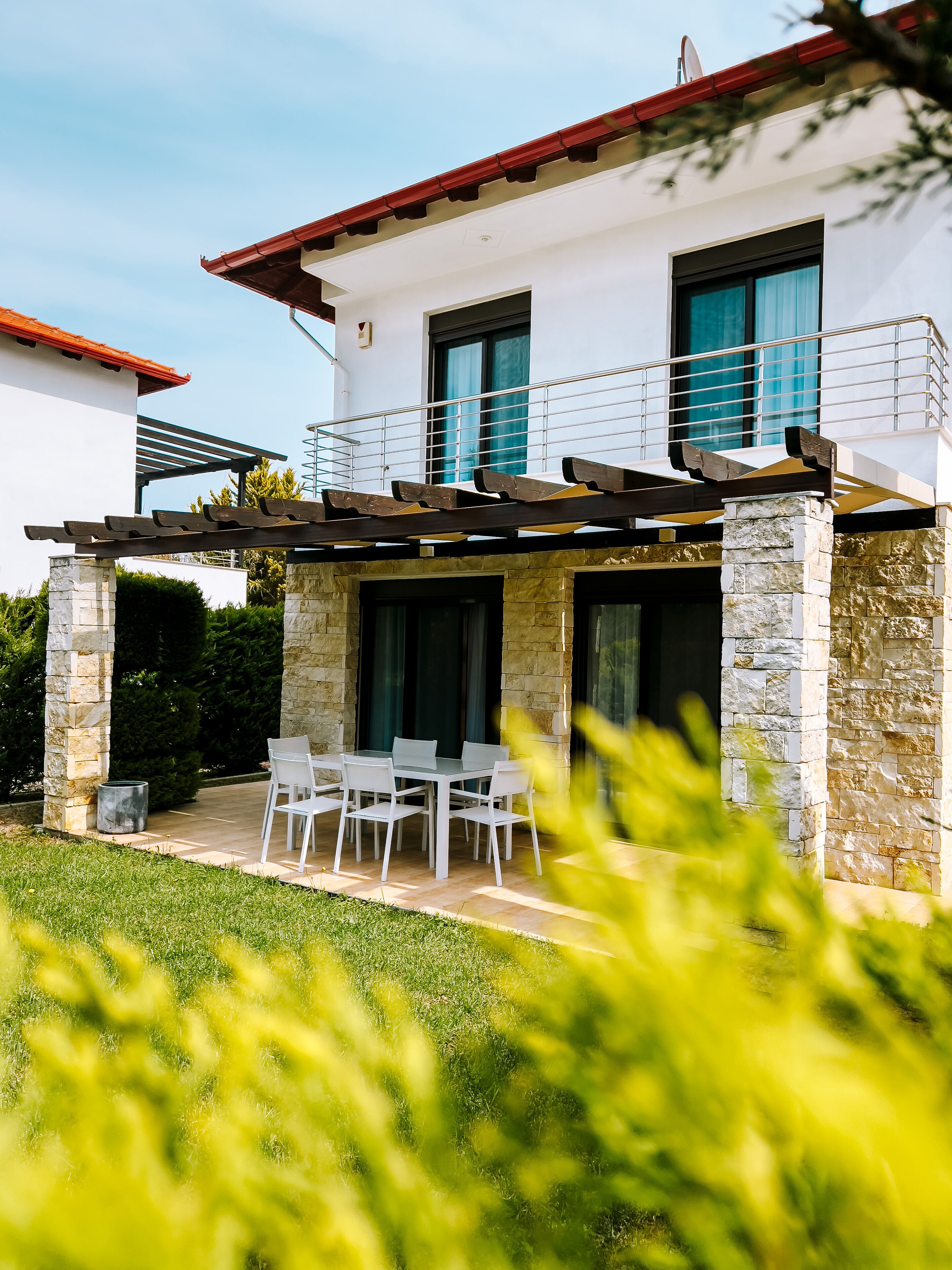 Uma casa moderna de dois andares com fachada de pedra e branca, uma varanda com grades de metal e um pátio com móveis de exterior.
