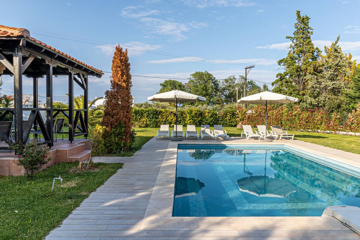 A rectangular swimming pool with a tiled deck, surrounded by lounge chairs and umbrellas, and a pergola structure to the left.